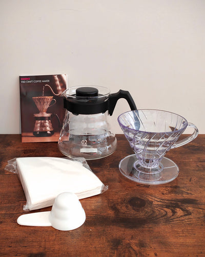 Coffee brewing equipment including a glass carafe, clear plastic filter, and white scoop on a wooden surface.